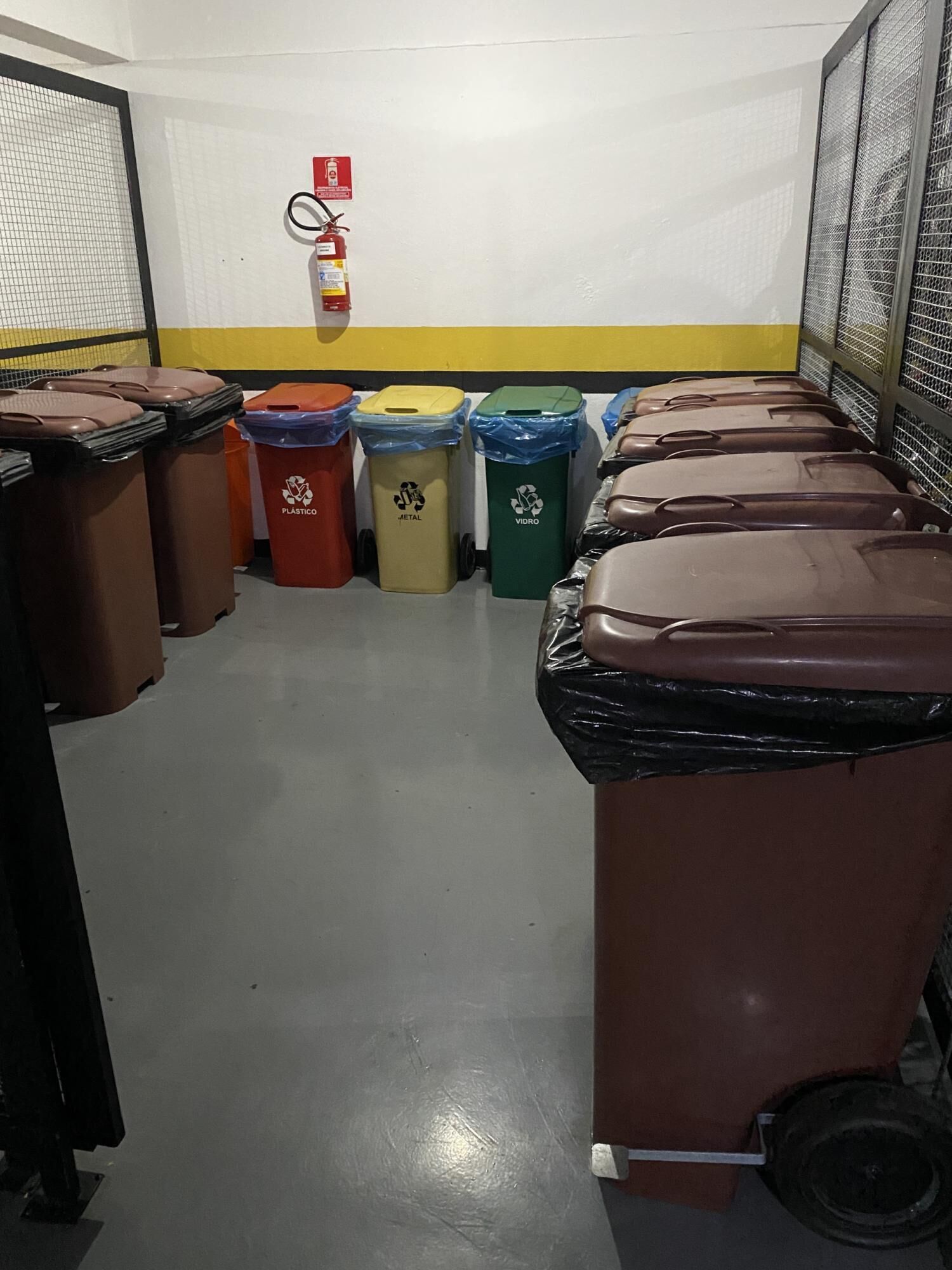 A row of trash bins in a room with a yellow stripe on the wall and a fire extinguisher mounted on the wall.