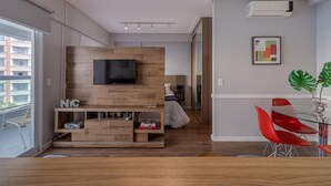 Un salon moderne avec un téléviseur à écran plat fixé sur un mur en bois, un coin repas avec des chaises rouges et une grande fenêtre donnant sur des immeubles.