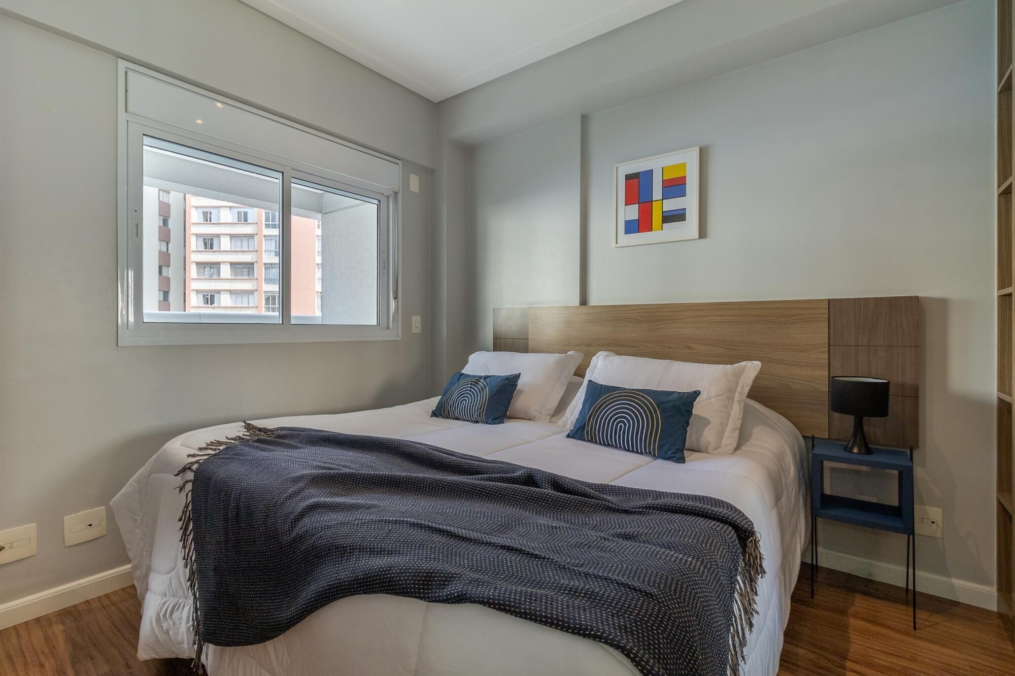 A bedroom with a large bed, a window with a view of buildings, a bedside table with a lamp, and a colorful abstract painting on the wall.