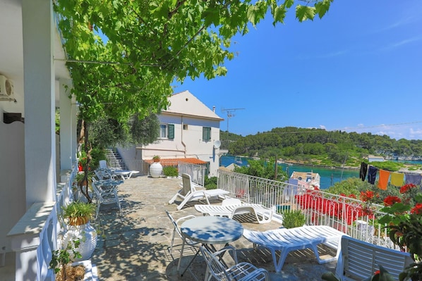 A terrace with outdoor seating, a white building, and a view of a body of water and greenery in the background.