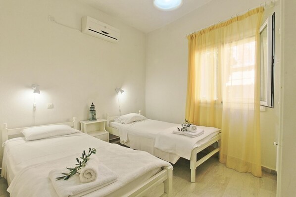 A hotel room with two beds, a bedside table with a lamp, an air conditioning unit, and a window with yellow curtains.