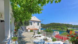 Eine Terrasse mit Sitzgelegenheit im Freien, ein weißes Gebäude und Blick auf ein Gewässer sowie Grünflächen.