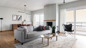 Una sala moderna con un sofá gris, una mesita de centro y una silla negra. Hay una chimenea, una zona de comedor con una mesa y sillas, y ventanas amplias que ofrecen vistas.