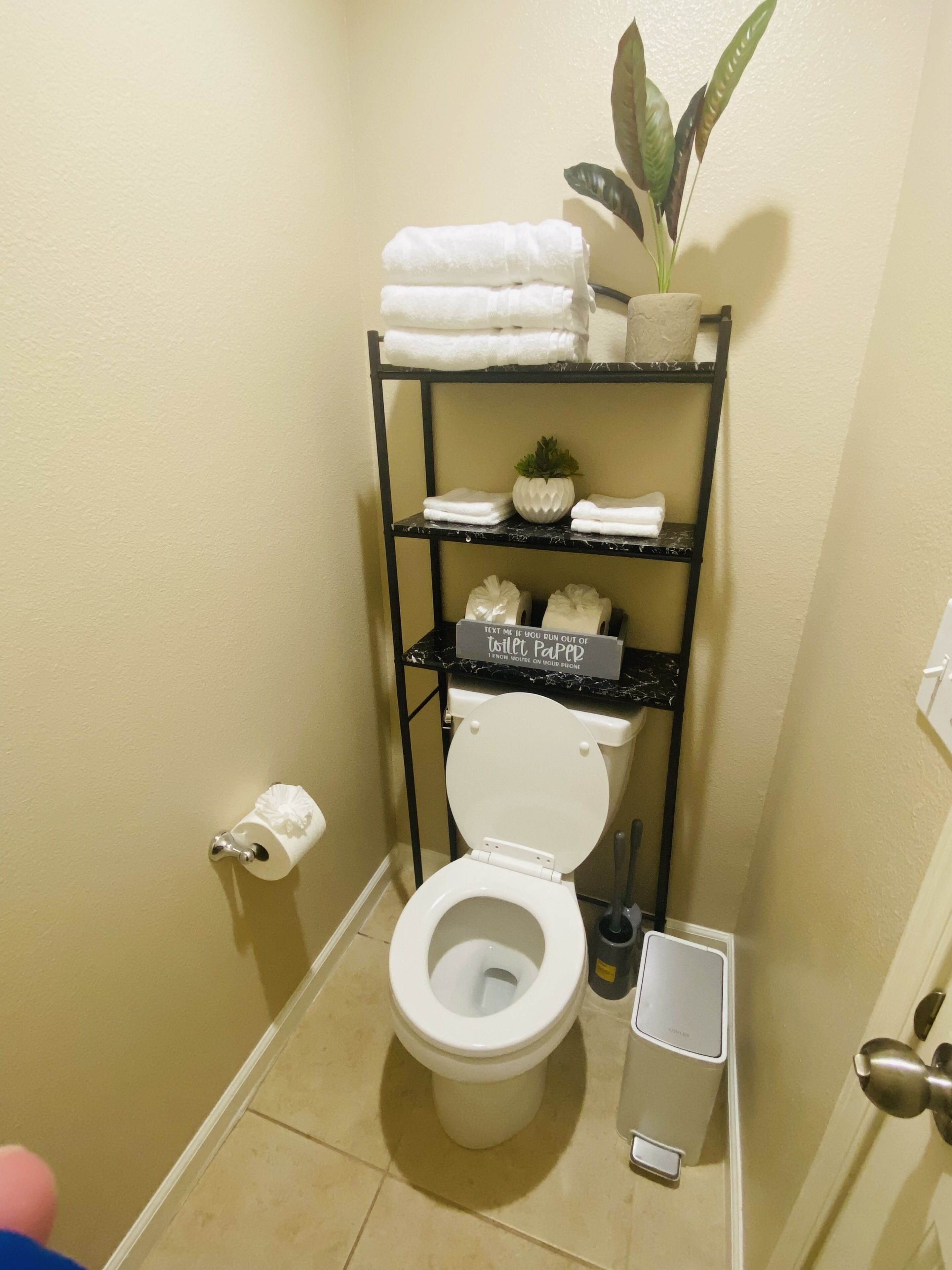 A compact bathroom with a white toilet, a shelf with folded towels, a potted plant, and a sign about toilet paper.