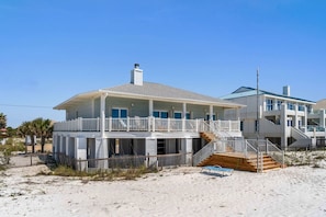 A two-story beachfront house with a balcony, a staircase, and a sandy beach in the background.