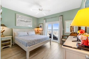 Et soveværelse med seng, ventilator, natbord med lampe, vindue med gardiner og havudsigt.