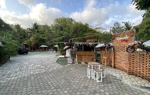 A courtyard with a brick wall, a sign for 'Kayuwata Cottage', outdoor seating, and a motorcycle parked against the wall. - Kayu Watu Cottage Gunung Kidul (Tepus)