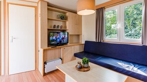 Een ruimtelijk ingerichte leefruimte met een televisie op een houten plank, een blauwe bank, een houten tafeltje met een plant en een raam met gordijnen.