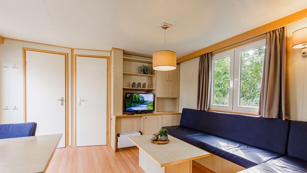 Een knus woongedeelte met een bank, een televisie op een plank, een tafel en een raam met gordijnen.