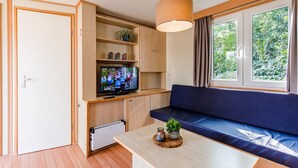 Ein kompakter Wohnbereich mit Fernseher, Holzschränken, einem blauen Sofa, einem Holztisch und einem Fenster mit Blick auf Grünflächen.