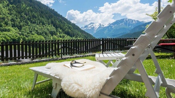 Een comfortabele lounge stoel met een zachte witte kussens, een boek en een zonnebril, gelegen op een gazon met een zwarte omheining en uitzicht op een berg.