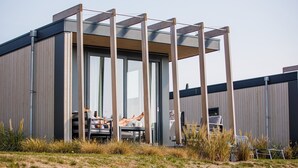 Un edificio moderno con grandes ventanales y vigas de madera. Cuenta con zonas de estar al aire libre, mesas y sillas.