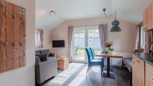 A modern living room with a sofa, a dining table with chairs, a TV mounted on the wall, and a large window with curtains. - Covered Terrace Chalet at Leukermeer (Well)