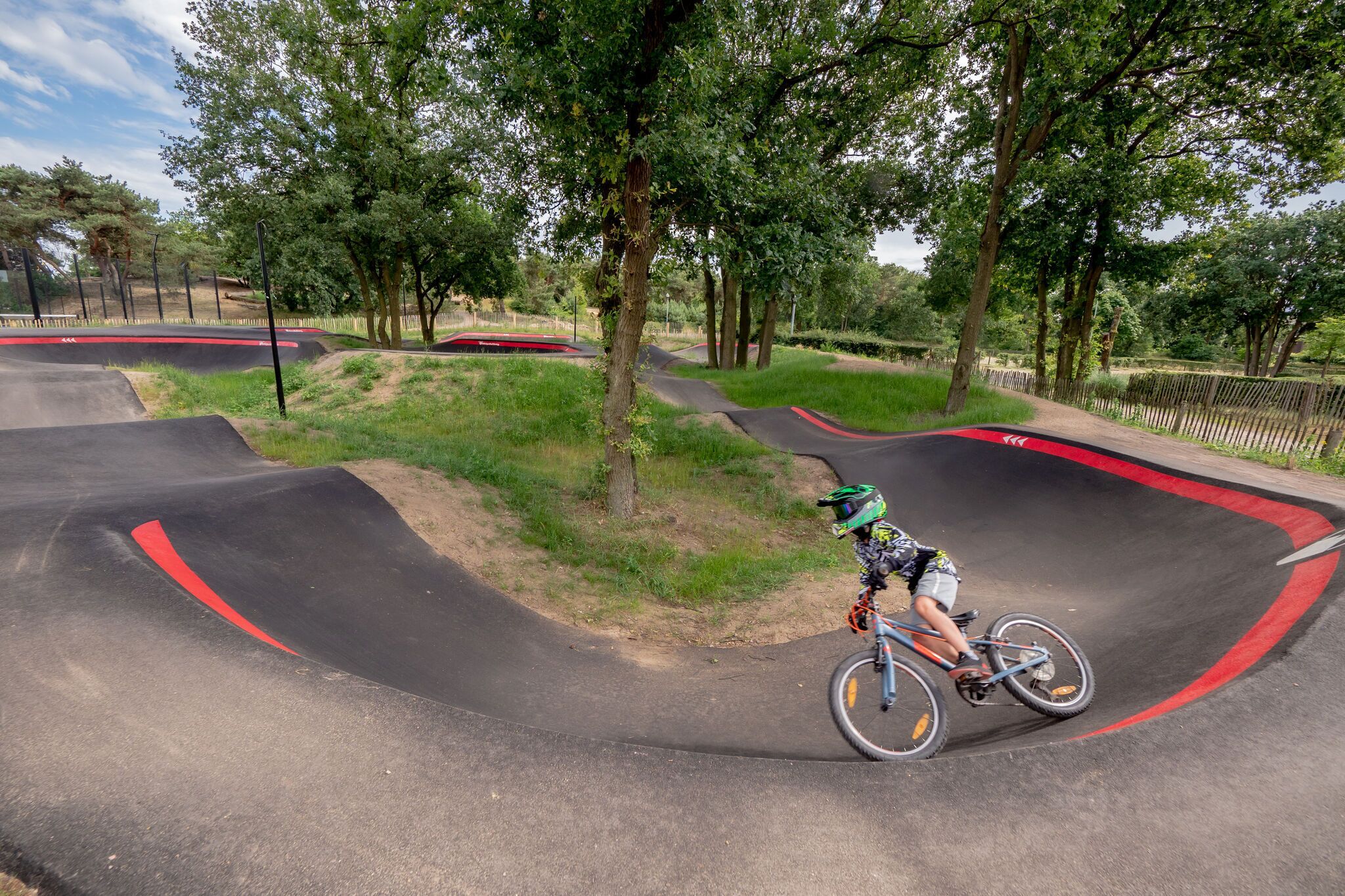 A person riding a bicycle on a curved track with red markings, surrounded by trees and grassy areas.