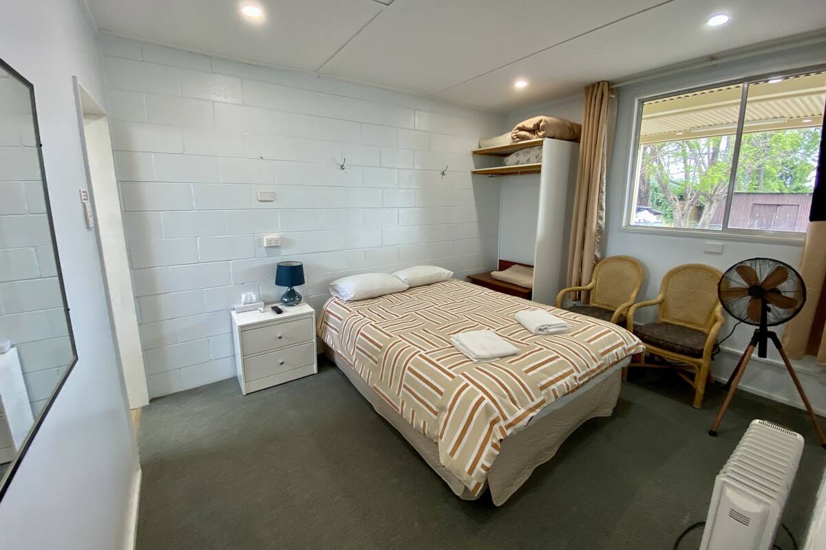 Une chambre à coucher avec un lit, une table de chevet, une chaise, un ventilateur et une fenêtre avec des rideaux.