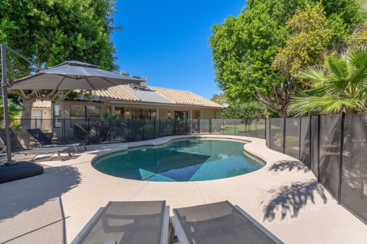 A swimming pool with a curved edge, surrounded by a fence, and shaded by a large umbrella.