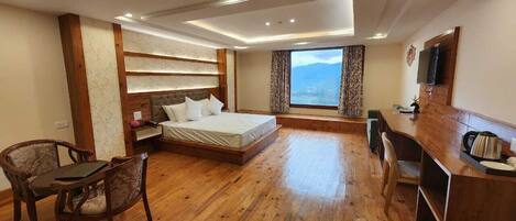 Una habitación de hotel con piso de madera, una cama con ropa blanca, un escritorio con televisor y un amplio ventanal con vistas a las montañas.