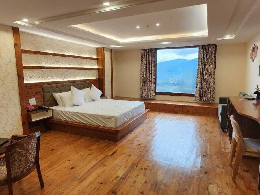 Una habitación de hotel con piso de madera, una cama con ropa blanca, un escritorio con televisor y un amplio ventanal con vistas a las montañas.