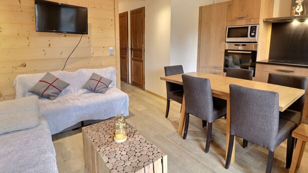 Una sala de estar acogedora con un sofá, una mesa de centro y un televisor montado en la pared. Hay una zona de comedor con una mesa y sillas. La cocina incluye un microondas y un horno.