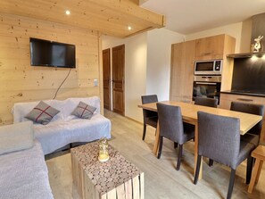 A cozy living room with a sofa, a coffee table, and a TV mounted on the wall. There is a dining area with a table and chairs. The kitchen area includes a microwave and oven.