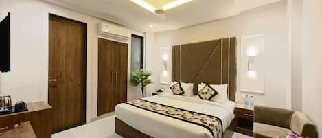 A modern hotel room with a large bed, a bedside table, a chair, a small table, a plant, and a ceiling light fixture.