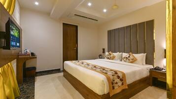 A hotel room with a large bed, a desk with a TV, a wooden headboard, and a bedside table with a lamp.