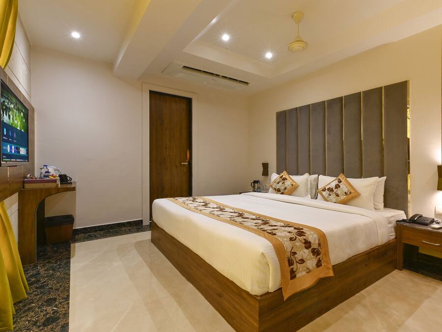 A hotel room with a large bed, a desk with a TV, a wooden headboard, and a bedside table with a lamp.