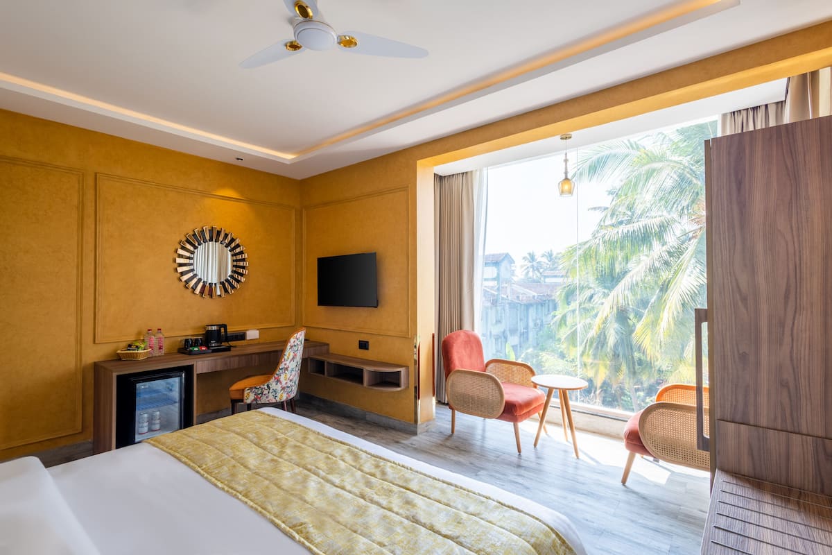 Quarto de hotel com uma cama, uma mesa com televisão, um mini frigorífico, uma cadeira e vista para palmeiras.