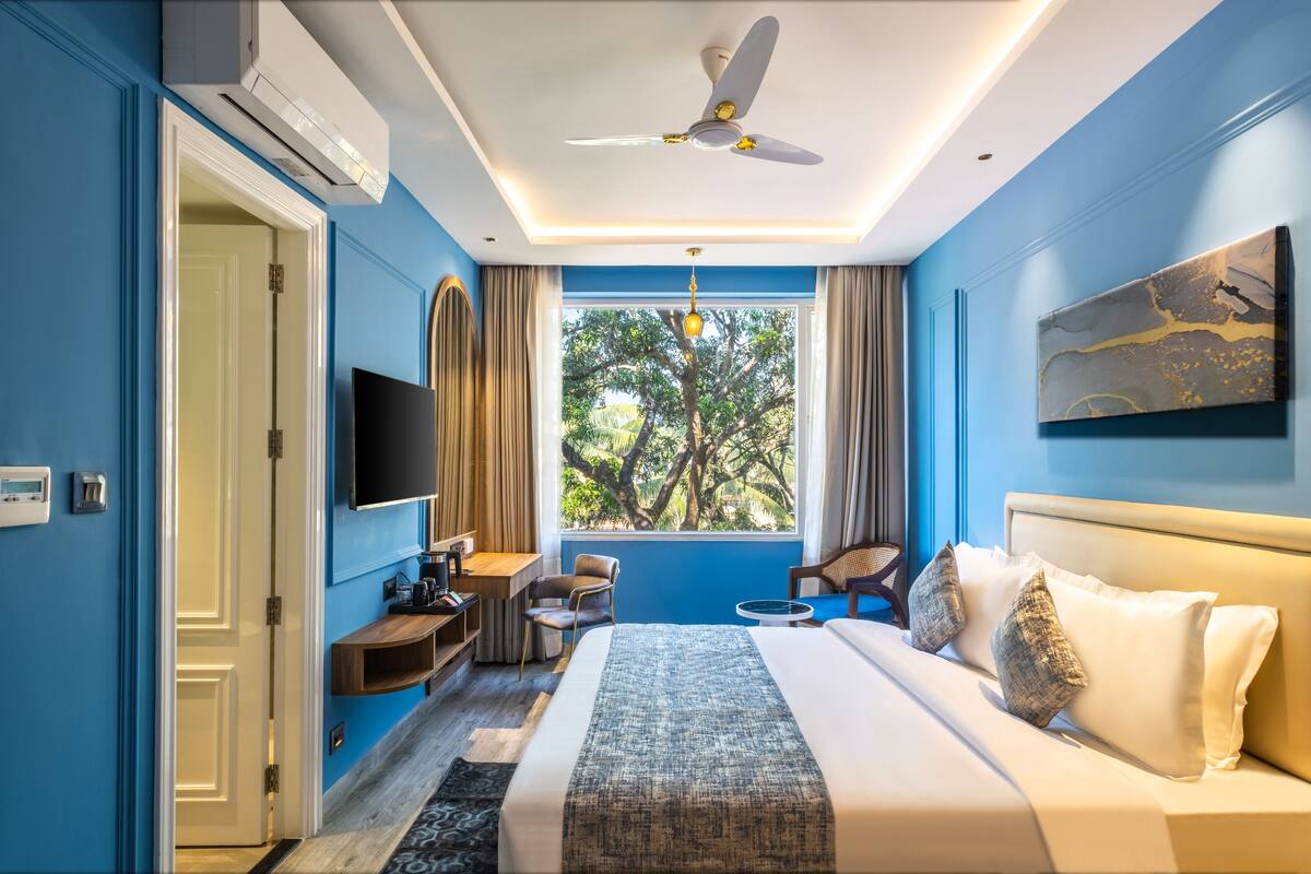 Quarto de hotel com uma cama grande, televisão, escrivaninha e janela com vista para árvores.