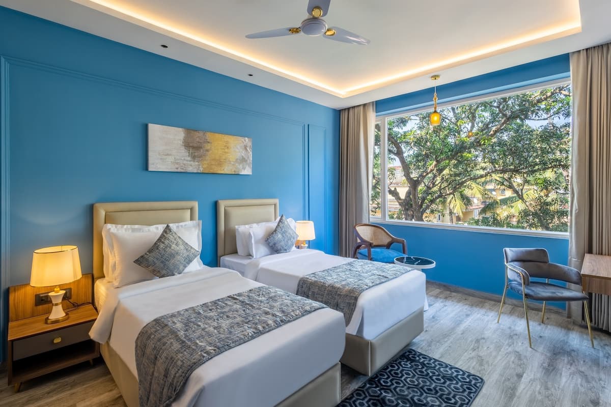 Quarto de hotel com duas camas, um ventilador de teto, uma janela com vista para árvores e uma mesa pequena com uma cadeira.