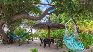 Un hamac sous un arbre, une table en bois avec des chaises et un parasol en chaume, le tout dans un cadre tropical.