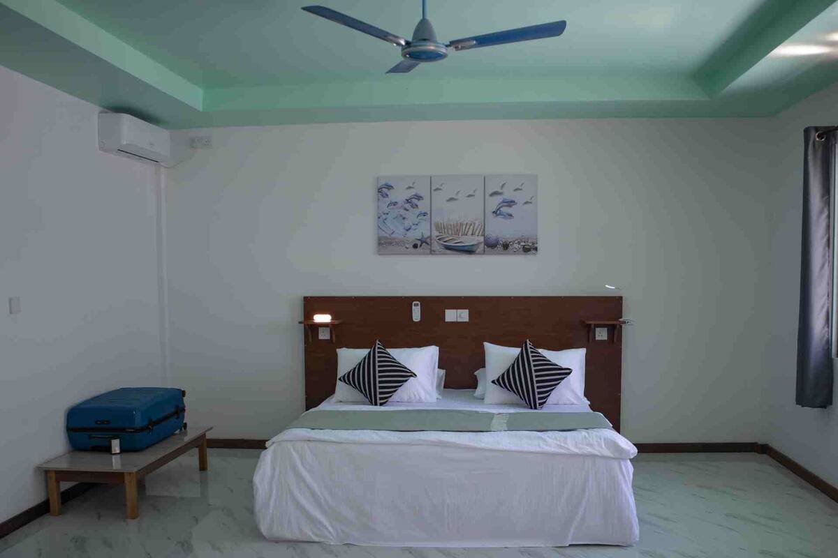 Ein Schlafzimmer mit einem Bett, einem Deckenventilator, einem blauen Koffer und Bildern an der Wand.