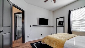 Ein Schlafzimmer mit Bett, einem an der Wand montierten Fernseher, einem Spiegel und einem Fenster mit Jalousien.
