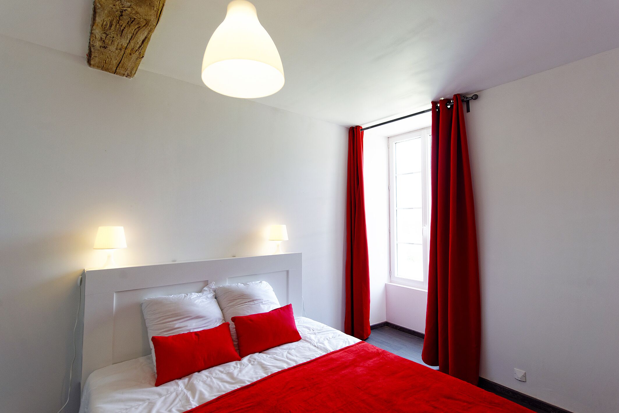 Een slaapkamer met een wit hoofdeinde, twee bedlampen, een rode sprei en rode gordijnen.