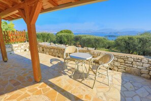 Relax in this serene outdoor seating area with a stone floor and wall, surrounded by a wooden pergola and offering a scenic view of a body of water and mountains.