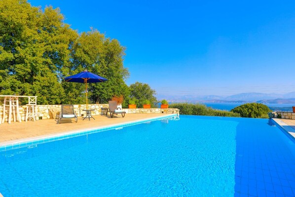 Une piscine d’un bleu éclatant, dotée de chaises longues et d’un parasol, entourée de murs de pierre et de verdure, et offrant une vue sur des montagnes lointaines.