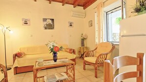 A living room with a sofa, armchairs, a coffee table, and a dining table. There are wall-mounted air conditioning units, a window with curtains, and a refrigerator. - Dina Beach Front Apartment (Corfu)