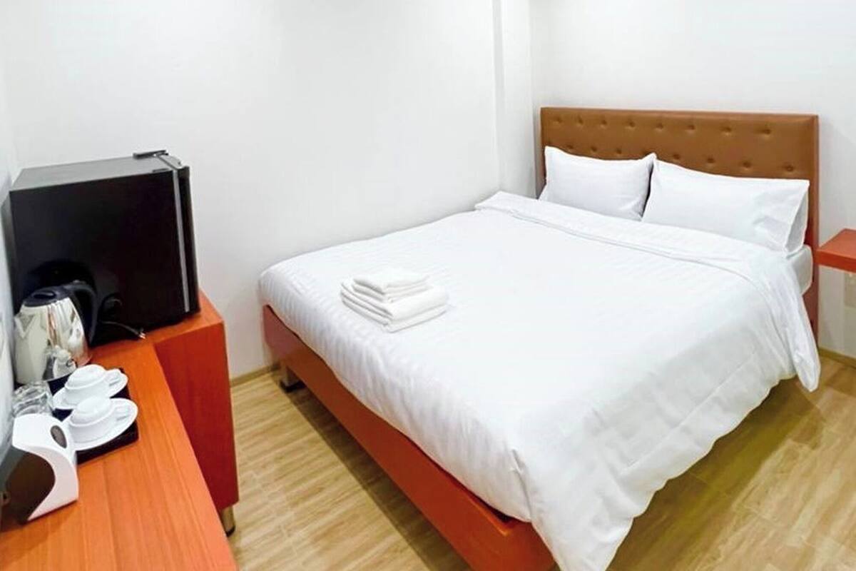 A hotel room with a bed, a wooden nightstand, a desk with a computer and a coffee maker, and a wall-mounted air conditioner.