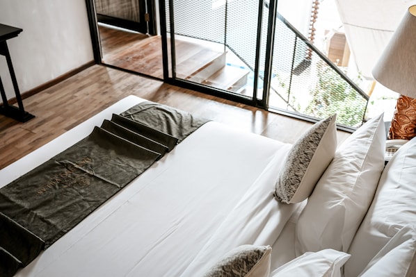 Una cama bien hecha con sábanas blancas y una manta marrón oscuro, una lámpara con pantalla estampada, y vistas a un balcón con barandilla de metal.