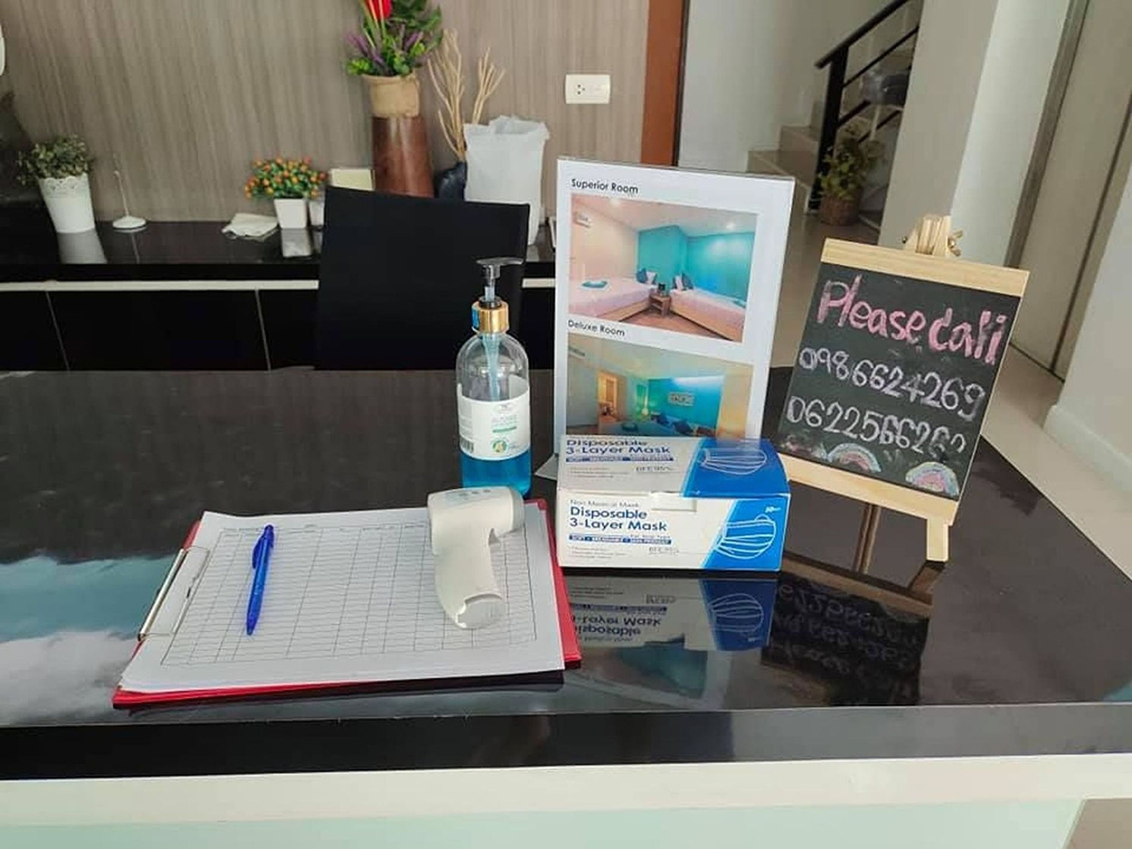 A hotel reception desk with a glass partition, a notepad, a pen, a disposable face mask, a sanitizer bottle, and a sign for the Superior Room.