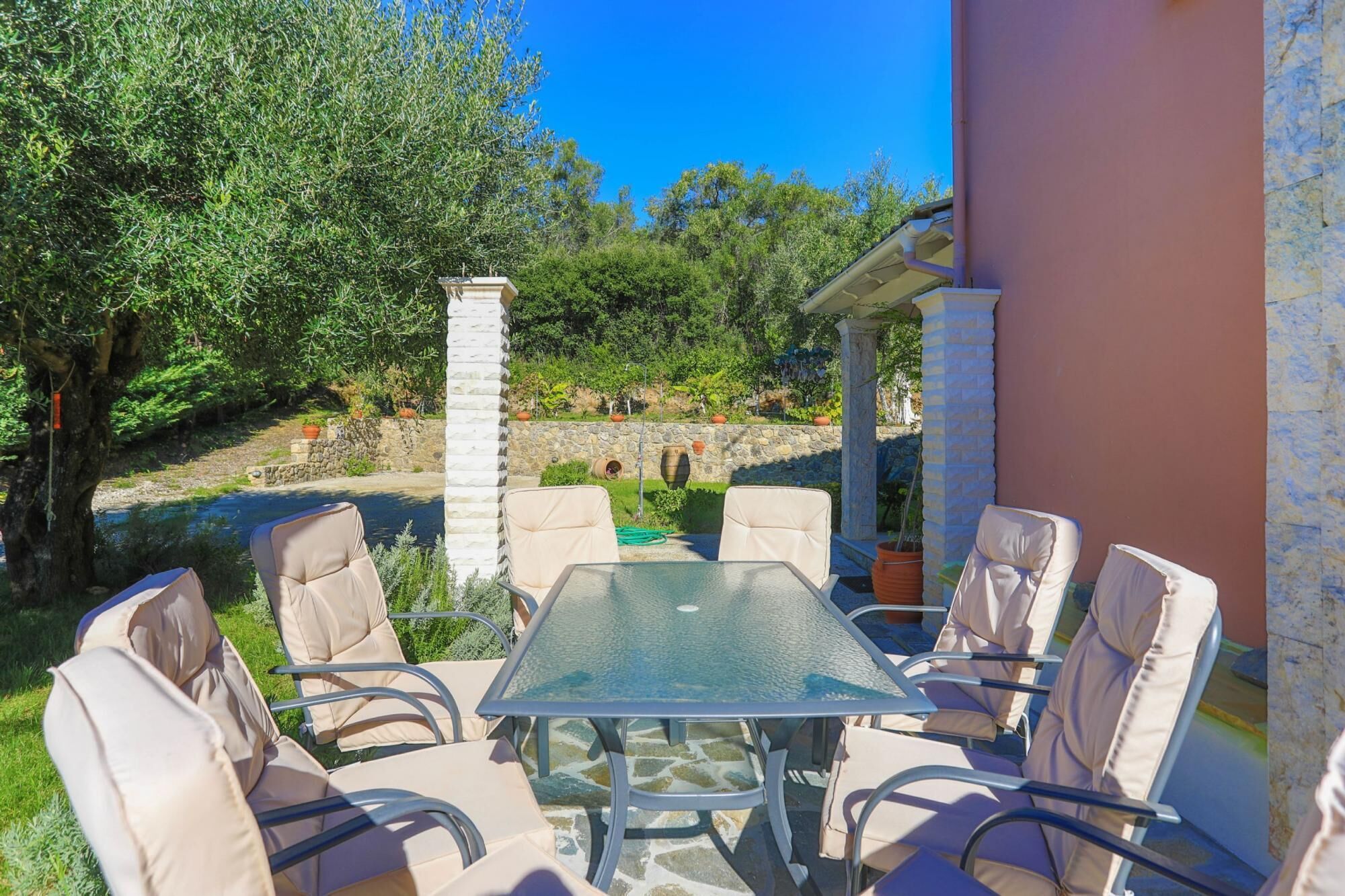 An outdoor patio with a glass table and six beige cushioned chairs.