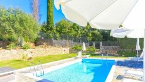 A swimming pool with clear blue water, surrounded by stone tiles and a stone wall. There are white lounge chairs and umbrellas around the pool. - Eleonas in Kato Korakiana (Corfu)