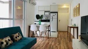 A compact living space with a teal sofa, a dining table with chairs, a kitchen area with a refrigerator, and a TV on a stand. - Iquali013 Akile Homes Apto (São Paulo)