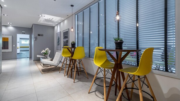 A modern dining area with a table and four yellow chairs, large windows with blinds, and a white sofa in the background. - Iquali013 Akile Homes Apto (São Paulo)