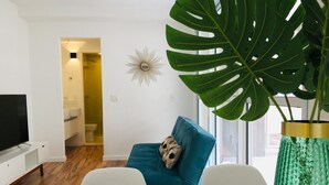 A modern living room with a TV, a blue sofa, a large green plant, and a white wall with a sunburst decoration. - Iquali013 Akile Homes Apto (São Paulo)