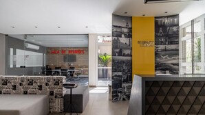 A modern lobby with a reception desk, a seating area with patterned cushions, and a wall with black and white photographs. - Iquali013 Akile Homes Apto (São Paulo)