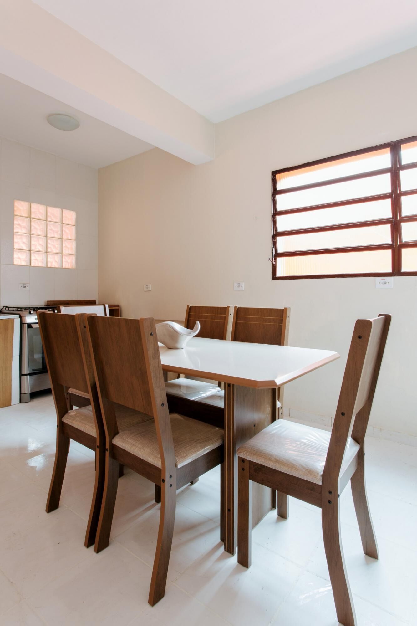 A dining area with a wooden table and chairs, a window with blinds, and a kitchen in the background.