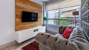 Een moderne woonkamer met een flatscreen televisie bevestigd aan een houten paneelwand, een grijze bank met een rood kussen, en een raam met uitzicht op gebouwen en groen.
