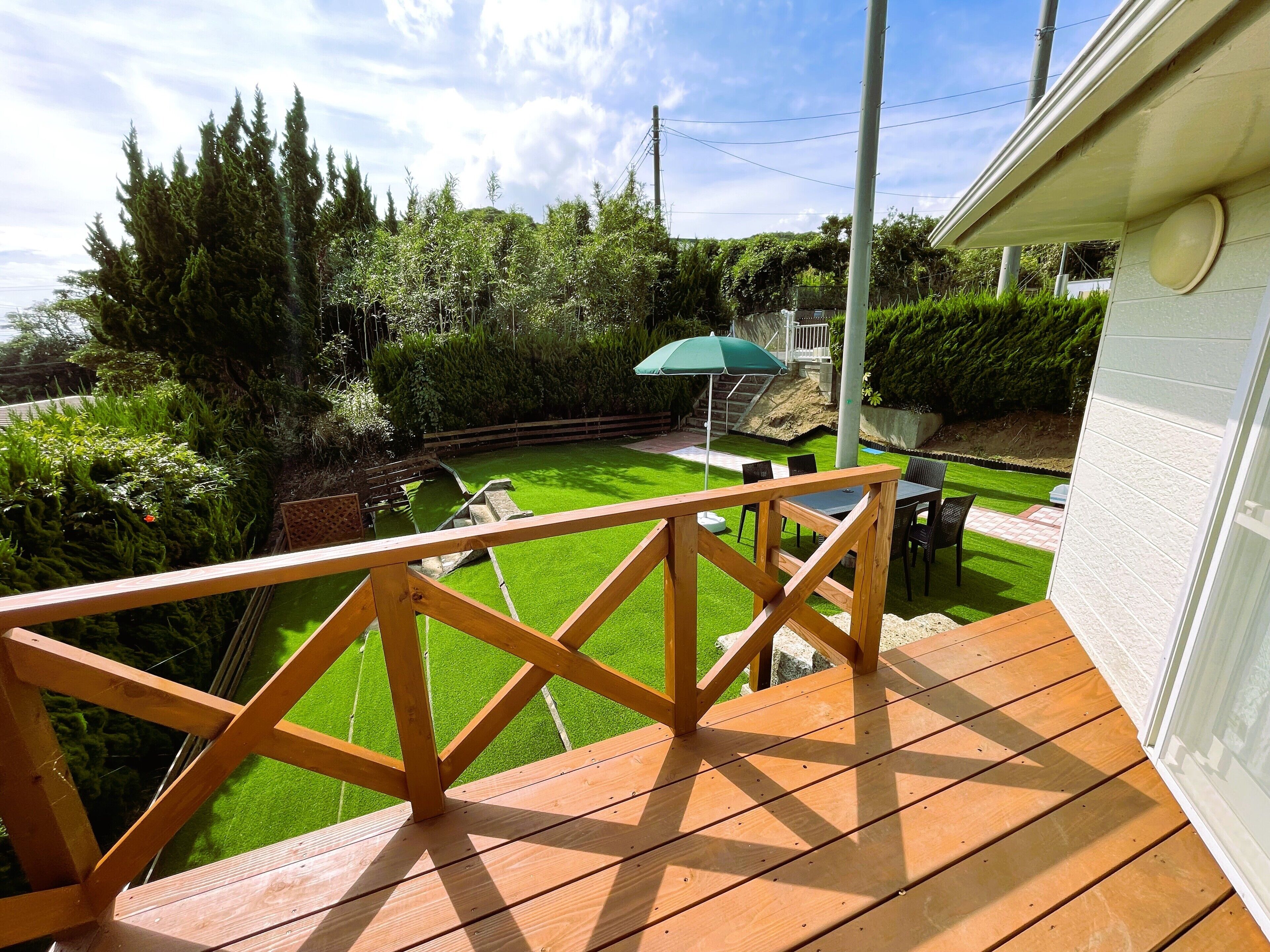 Una terraza de madera con barandilla, con vistas a un patio que tiene césped, una mesa, sillas y un parasol verde.