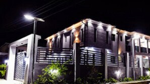 A modern building with a dark facade, illuminated by streetlights, featuring a prominent entrance with a metal gate and a small overhang above the door. - Tito Empire (Lekki)
